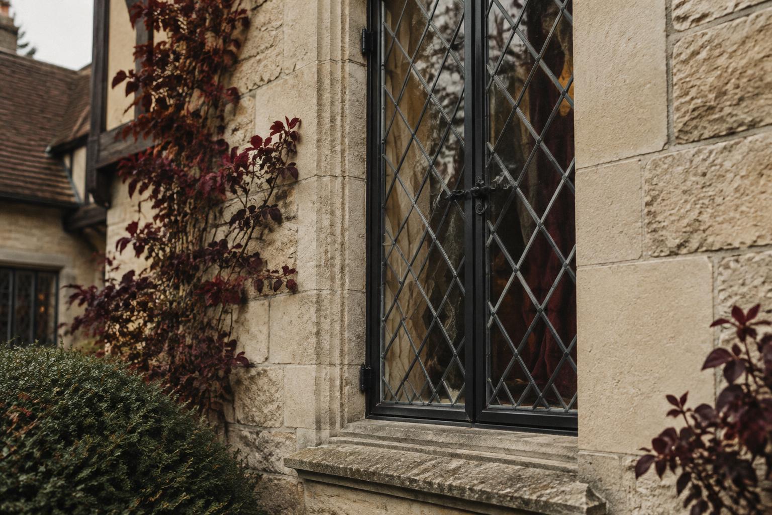 Black steel casement window with diamond leaded glass in a limestone wall of a Tudor Revival house, framed by burgundy foliage.